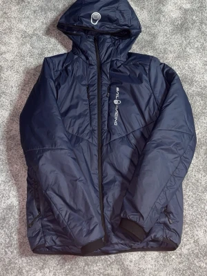 Mörkblå Sail Racing vindjacka S - Säljer en mörkblå vindjacka från Sail Racing med huva och hel dragkedja. Jackan har två sidofickor med dragkedja, justerbara muddar och logga på bröstet. Materialet är lätt och vindtätt, perfekt för blåsiga dagar. Enkel och stilren design.