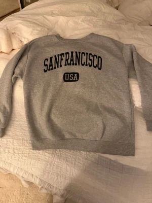 Grå sweatshirt med SANFRANCISCO-tryck - Säljer en grå sweatshirt med svart SANFRANCISCO USA-tryck. Tröjan har rund halsringning och långa ärmar. 