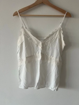 Vit spetsdetaljerad linnetopp från Vila - Säljer en vit linnetopp från Vila i storlek 36. Toppen har tunna axelband, v-ringning och fina spetsdetaljer längs halsringningen och tvärs över bröstet. Materialet är mjukt och har en lätt glans, vilket ger en lyxig känsla.