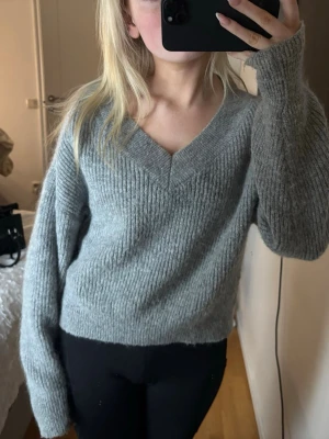Grå stickad v-ringad tröja - Mysig grå stickad tröja med v-ringning och lång ärm. Tröjan har en relaxed passform och ribbad struktur som ger en skön och avslappnad look. Perfekt att slänga på sig under kyliga dagar och funkar till allt.