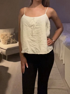 Vit linnetopp med smala axelband - Säljer en enkel och fräsch vit linnetopp med smala axelband och liten knytning framtill. Toppen är ärmlös och har en lös passform, perfekt för varma dagar.