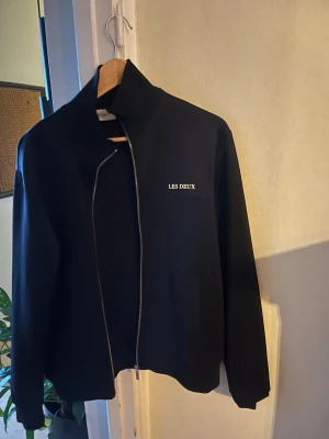 Svart zip hoodie från Les Deux - Säljer en stilren svart bomberjacka från Les Deux med diskret logga på bröstet och hel dragkedja framtill. Jackan har hög krage och raka ärmar, perfekt för dig som gillar minimalistisk design.