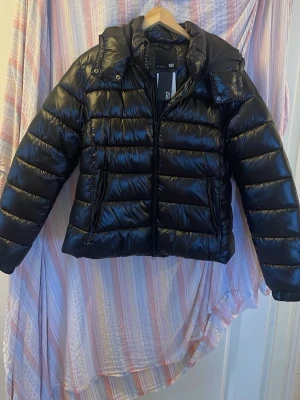 Svart pufferjacka från 157, XL - Säljer en svart, glansig pufferjacka från 157 i storlek XL. Jackan har hög krage, dragkedja framtill och två sidofickor med dragkedja. Perfekt för kalla dagar och riktigt snygg till streetwear-looken.