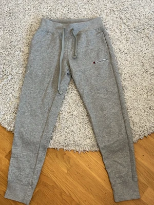 Grå mjukisbyxor från Champion XS - Grå mjukisbyxor från Champion i storlek XS. Byxorna har dragsko i midjan, ribbade muddar vid bensluten och Champion-logga broderad på ena benet. Tillverkade i en mjuk bomulls- och polyestermix för maximal komfort.
