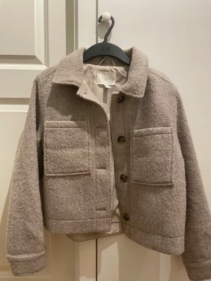 Beige kappa från H&M XS - Säljer en beige kappa från H&M i storlek XS. Jackan har två stora fickor framtill, klassisk krage och stängs med knappar. Den är i ett mjukt och  material som ger en chill och trendig look. Perfekt för lager-på-lager under höst och vinter.
