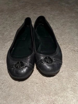 Svarta ballerinaskor från Ralph Lauren - Klassiska svarta ballerinaskor från Ralph Lauren med rund tå och dekorativt RLL-emblem i metall framtill. Skorna är tillverkade i skinn och har en platt sula för skön komfort. Perfekta till jeans eller kjol för en clean och stilren look. Skriv ifall ni vill ha fler bilder eller har några frågor!❣️Pris går att diskutera 🥰
