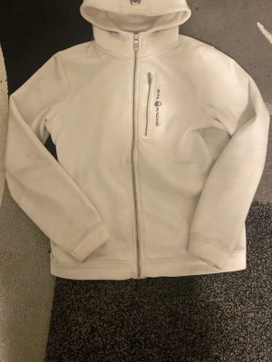 Vit fleecejacka från Sail Racing - Säljer en vit fleecejacka från Sail Racing med huva och dragkedja framtill. Jackan har en bröstficka med dragkedja och logga på bröstet. Perfekt för kyliga dagar och enkel att matcha med allt.
