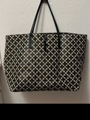 Svart och beige shoppingväska By Malene Birger - Stor shoppingväska från By Malene Birger i svart och beige med ikoniskt grafiskt mönster. Väskan har långa svarta handtag och rymligt beige innerfoder. Tillverkad i slitstarkt syntetmaterial och stängs med magnetknapp. Perfekt för dig som gillar stilrena detaljer. Sliten på handtagen tagit bild på alla skador annars fint skick