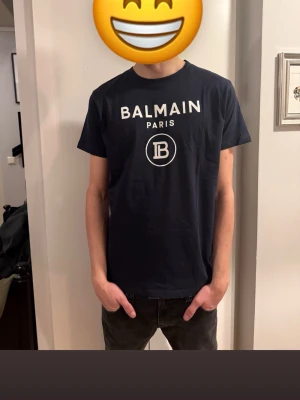 Mörkblå Balmain Paris t-shirt - Snygg mörkblå t-shirt från Balmain Paris med vit logga och tryck framtill. Klassisk rund hals och korta ärmar. Tillverkad i mjuk bomull som känns skön mot huden. Perfekt för dig som vill ha en clean och stilren look.