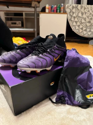 Nike Air fotbollsskor lila/svart - Säljer ett par riktigt snygga Nike Air fotbollsskor i lila och svart med orange detaljer. Skorna har strumpliknande skaft, snörning och en cool sula med orange och lila inslag. Ovandelen är i syntetmaterial med vågiga svarta linjer för extra stil.