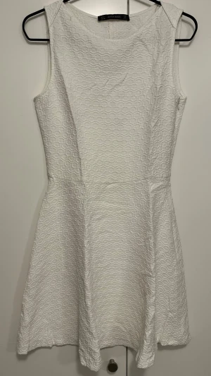 Vit kort klänning från Zara Basic - Supersnygg vit kort klänning från Zara Basic i strukturerat tyg med vågigt mönster. Ärmlös modell med rund halsringning och figurnära passform upptill som går ut i en lätt utställd kjol.