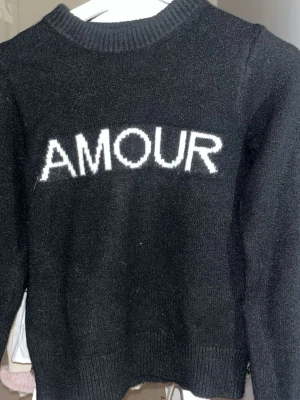 Svart stickad tröja med AMOUR-text - Säljer en svart stickad tröja med vit text 'AMOUR' framtill. Perfekt för dig som gillar enkel men snygg stil med ett statement. 🩶
