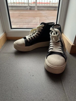 Svarta high-top sneakers från Good News - Säljer ett par svarta high-top sneakers från Good News med vit platt sula och rund tå. Skorna har vita snören, kontrastsömmar och vit tå. Materialet är canvas och detaljerna ger en cool och stilren vibe. Perfekt för dig som gillar chunky sneakers med retrokänsla.
