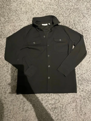 Svart overshirt från Only & Sons - En svart overshirt är ett stilrent och mångsidigt plagg som passar perfekt året runt. Tillverkad i ett bekvämt och slitstarkt material med en tidlös design som enkelt kan bäras både som skjorta och som lätt jacka. Den svarta färgen ger ett modernt och sofistikerat uttryck, medan den avslappnade passformen gör den lika snygg till vardags som till mer uppklädda tillfällen. Perfekt att kombinera med jeans, chinos eller över en t-shirt för en trendmedveten look.
