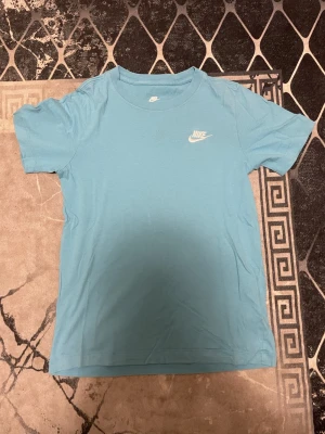 Blå Nike t-shirt i bomull - Säljer en ljusblå t-shirt från Nike med klassisk logga broderad i vitt på bröstet. T-shirten har rund halsringning och korta ärmar. Tillverkad i mjuk bomull för skön känsla och enkel stil.