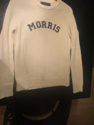 Vit stickad tröja från Morris - Snygg vit stickad tröja från Morris med mörkblå text framtill. Klassisk rund halsringning och långa ärmar. Perfekt till jeans eller chinos för en clean och stilren look. Den har några defekter på underarmen, som du kan se på bild 5 men de går att ta bort med rengöring, men som sagt så syns de inte mycket eftersom de är på underarmen 