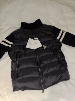 Moncler dunjacka – XS – Nyskick med tags - Säljer en exklusiv Moncler dunjacka i storlek XS i svart. Jackan har den ikoniska Moncler-designen med stickade ärmar med vita ränder och logopatch på armen, vilket ger en sportig men lyxig look.  ✔ Original Moncler ✔ Helt ny – med alla tags kvar ✔ Lätt, varm och bekväm ✔ Dragkedjor i metall med Moncler-logga ✔ Perfekt för höst, vinter och vår  Jackan är aldrig använd och i perfekt skick. Nypris är betydligt högre – perfekt tillfälle att fynda en  Moncler till bra pris.  Skickas snabbt
