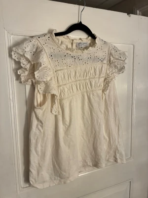 Vit blus med spets från Holly & Whyte - Säljer en superfin vit blus från Holly & Whyte by Lindex. Blusen har broderade spetsdetaljer och volang vid ärmarna samt runt halsen. Den är kortärmad och har ett romantiskt, luftigt uttryck med blommigt hålmönster upptill. Perfekt till våren eller sommaren!