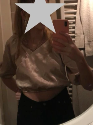 Champagnefärgad omlottopp från BikBok - Säljer en champagnefärgad omlottopp från BikBok i glansigt material. Toppen har ballongärmar, resår i midjan och vid ärmsluten samt en croppad passform. 