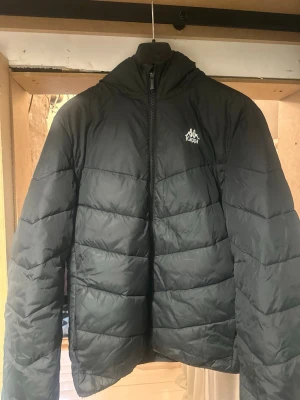 Svart Kappa pufferjacka  - Säljer en svart pufferjacka från Kappa med huva och dragkedja. Jackan har Kappa-logga på bröstet och snygga loggor längs huvan. Använd en vinter. Hör av dig vid frågor!