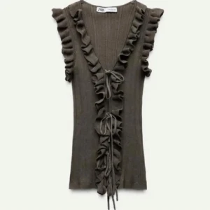 Olivgrön ribbad topp med volang Zara - Supersnygg olivgrön ribbad topp från Zara med volangdetaljer längs axlar och v-ringning. Toppen har snörning framtill och är ärmlös, vilket ger en trendig och cool look. Perfekt att styla med jeans eller kjol för en chic outfit.