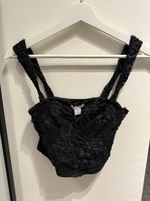 Svart spetsig korsettopp med blommor - Säljer en svart korsettopp från Missguided med blommiga spetsdetaljer och breda axelband. Toppen har en liten v-ringning framtill och är croppad, perfekt till höga jeans eller kjol. Prislapp är fortfarande 