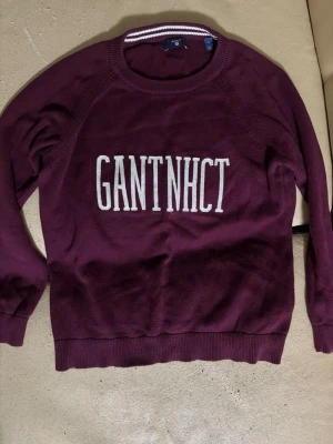Vinröd stickad tröja från Gant - Säljer en vinröd stickad tröja från Gant med vit text 'GANT NHCT' framtill. Tröjan har rund halsringning med vit randig detalj och långa ärmar. Perfekt för dig som gillar klassisk college-stil.
