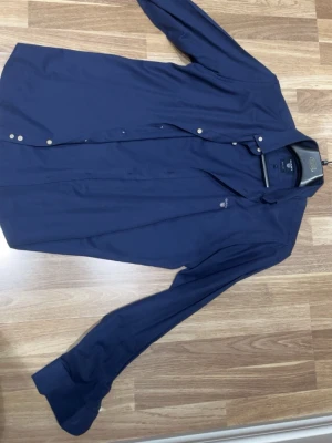 Marinblå skjorta från Gant Slim Fit - Säljer en marinblå skjorta från Gant i storlek L men sitter som S/m med slim fit-passform. Skjortan har klassisk krage, långa ärmar och vita knappar framtill. Perfekt för dig som gillar stilrena och tidlösa plagg. Aldrig använd fick i present 