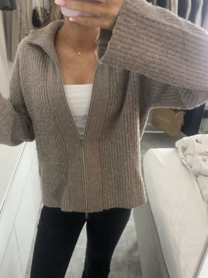 Beige ribbad kofta med dragkedja - Mysig beige ribbad kofta med lång ärm och hög krage. Koftan har en tvåvägsdragkedja framtill och är stickad i ett mjukt material som känns riktigt skönt.🤎