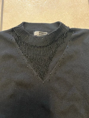 Svart stickad tröja AK Collection storlek S - Svart stickad tröja från AK Collection med lång ärm och ribbad hals. Snygga svarta pärlor pryder halsringningen i ett V-format mönster framtill. Perfekt för dig som gillar detaljer och vill ha en stilren look.