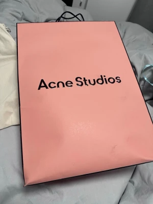 Brun axelväska från Acne Studios - Säljer en snygg brun axelväska från Acne Studios i slätt skinn. Väskan har stilrena handtag, diskret logga framtill och magnetknäppning. Insidan har ett band med Acne Studios-tryck. Perfekt för dig som gillar minimalistisk design och vill ha en trendig accessoar.