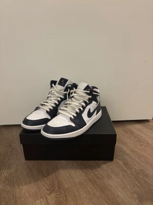 Nike Air Jordan 1 Mid Marinblå/Vit - Säljer ett par Nike Air Jordan 1 Mid i marinblått och vitt skinn. Klassisk siluett med snörning, rund tå och vit sula. Kommer med originalkartong och extra skosnören. Använda endast en gång