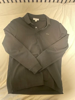 Svart pikétröja från Lacoste - Klassisk svart pikétröja från Lacoste med diskret krokodillogga på bröstet. Tröjan har krage, två knappar framtill och långa ärmar. Regular fit-modell i mjuk bomull som passar perfekt till både jeans och chinos.