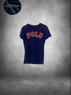 Blå Polo Ralph Lauren t-shirt - Snygg blå t-shirt från Polo Ralph Lauren med stor röd och vit POLO-text på bröstet. Modellen är kortärmad och har en klassisk rund hals. Passformen är custom fit och materialet är mjuk bomull, perfekt för en chill och stilren look.