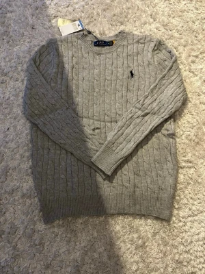 Grå kabelstickad tröja Polo Ralph Lauren - Snygg grå kabelstickad tröja från Polo Ralph Lauren i storlek M. Klassisk rund halsringning och ikoniska broderade loggan på bröstet. Perfekt till lager på lager och passar till allt. Mjuk och skön kvalitet i ullblandning.