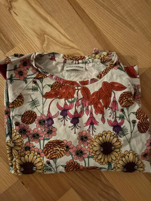 Blommig t-shirt från Mini Rodini - Säljer en färgglad t-shirt från Mini Rodini med ett stort blommigt mönster i rosa, lila, orange och gul på vit botten. T-shirten har rund hals och långa ärmar, perfekt för dig som gillar unika prints och lekfull stil.