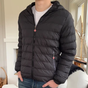 Svart pufferjacka från Colmar - Svart pufferjacka från Colmar Originals med huva och dragkedja framtill. Jackan har quiltad design, två sidofickor med dragkedja och en snygg logotyp på ärmen. Perfekt för kyliga dagar och har en clean, modern look.