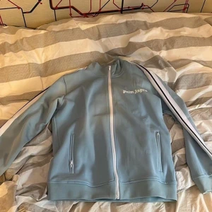 Palm Angels ljusblå zip hoodie - Snygg ljusblå hoodie från Palm Angels med vit dragkedja och vita ränder längs ärmarna. Tröjan har hög krage, två fickor med dragkedja och logotypen tryckt på bröstet. Perfekt för dig som gillar streetwear och vill sticka ut.