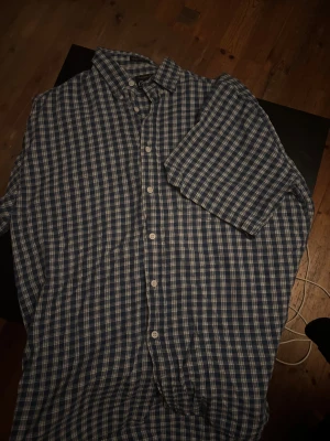 Blårutig skjorta från Cornwell M - Blårutig skjorta från Cornwell i storlek M, tillverkad i 100% bomull. Skjortan har korta ärmar, klassisk krage och knäppning framtill med vita knappar. Perfekt för dig som gillar en chill och tidlös stil med rutigt mönster.