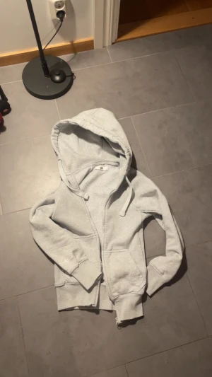 Ljusgrå flecce från russemerch  - Säljer en ljusgrå hoodie från Russemerch i storlek XXS. Den har dragkedja, två fickor fram och en stor huva med dragsnören. Insidan är mjukt fodrad för extra komfort. Perfekt för dig som gillar en enkel och clean stil.