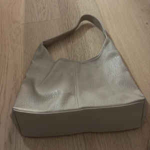 Beige axelväska i skinn - Snygg beige axelväska med stilren och mjuk design. Väskan har en bred axelrem och är tillverkad i skinn med lätt textur. Perfekt storlek för att få plats med allt du behöver och passar till många olika outfits.