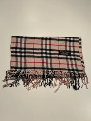 Burberry halsduk i klassiskt rutmönster - Snygg halsduk från Burberry i beige, svart, vitt och rött klassiskt rutmönster. Tillverkad i 100% lammull och har fransar i ändarna. Perfekt accessoar för att lyfta din outfit med en ikonisk brittisk touch.