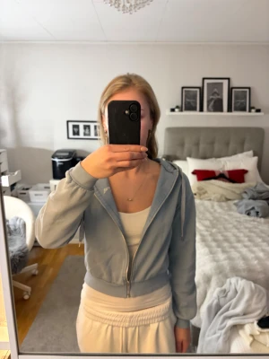 Ljusblå hoodie Beverly Hills - Snygg ljusblå hoodie med dragkedja och broderad Beverly Hills-text på bröstet. Modellen är croppad med ribbade muddar och har en klassisk huva med dragsko. Mjuk bomullsmix som är skön att ha på sig och passar perfekt till jeans eller mjukisbyxor. 