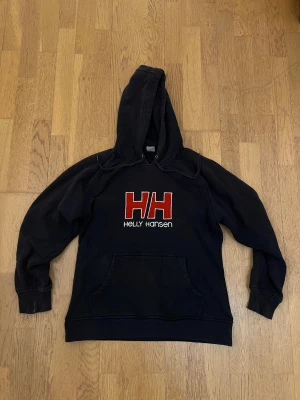 Svart hoodie från Helly Hansen - strl.L - Svart hoodie från Helly Hansen med stor röd HH-logga och vit text på bröstet. Tröjan har huva med snörning och en klassisk magficka. Materialet är mjuk bomull och polyester, perfekt för chill dagar. Snygg och enkel att matcha med jeans eller joggers.
