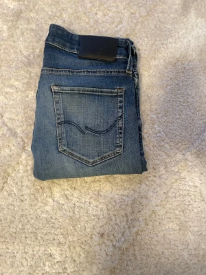 Jack n Jones jeans blå slim fit - Snygga blå Jack n Jones jeans med klassisk femficksdesign och svarta läderpatch bak. Slim fit med lätt tvättade detaljer.Perfekt passform och stilren look för dig som gillar jeans med lite attityd.