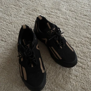 Svarta sneakers från EA7 Emporio Armani - Snygga svarta sneakers från EA7 Emporio Armani med beigea grafiska detaljer och coola mönster på sidorna. Skorna har snörning, rund tå och en chunky, mönstrad sula. Ovandelen är i textil och syntet, perfekt för en sportig och trendig look.