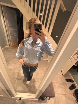 Ljusblå volangblus med knyt - Supersöt ljusblå blus med volanger längs axlarna och bröstet. Blusen har ett litet knyt i halsen och ballongärmar med resår vid handlederna. Croppad modell i tunt, krispigt tyg som ger en luftig känsla. Perfekt för sommarn och våren