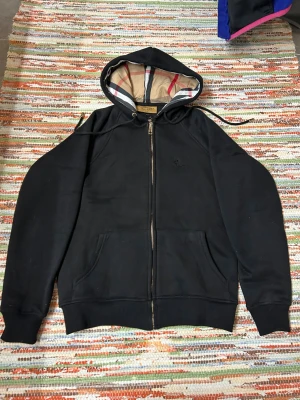 Svart Burberry hoodie med dragkedja - Svart hoodie från Burberry med klassiskt rutigt foder i huvan och guldfärgad dragkedja. Tröjan har broderad Burberry-logga på bröstet, känguruficka och justerbar huva. Tillverkad i mjuk bomull, perfekt för en chill och stilren look. Sitter VÄLDIGT cropped och tight. 