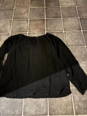 Svart blus med knytning från 157 - Svart blus i tunn, skrynklig bomull med v-ringning och snygg knytning fram. Långärmad modell med dekorativa spetsdetaljer längst ner. Perfekt för dig som gillar en chill och stilren look.