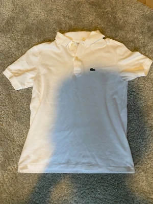 Vit pikétröja från Lacoste - Vit snygg pikétröja köpt direkt från Lacostes hemsida med klassisk krage, korta ärmar och knäppning framtill. Den har mycket fint skick.Tveka inte att skriva om ni har frågor!